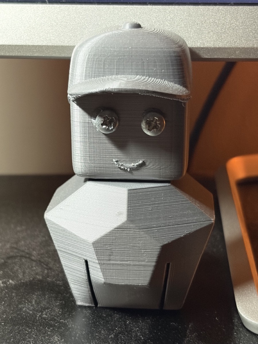 Two grey 3D-printed parts on a desk: a faceted angular head with a horizontal visor slot, and a rounded rectangular torso with two black Pozidriv screws through its face.